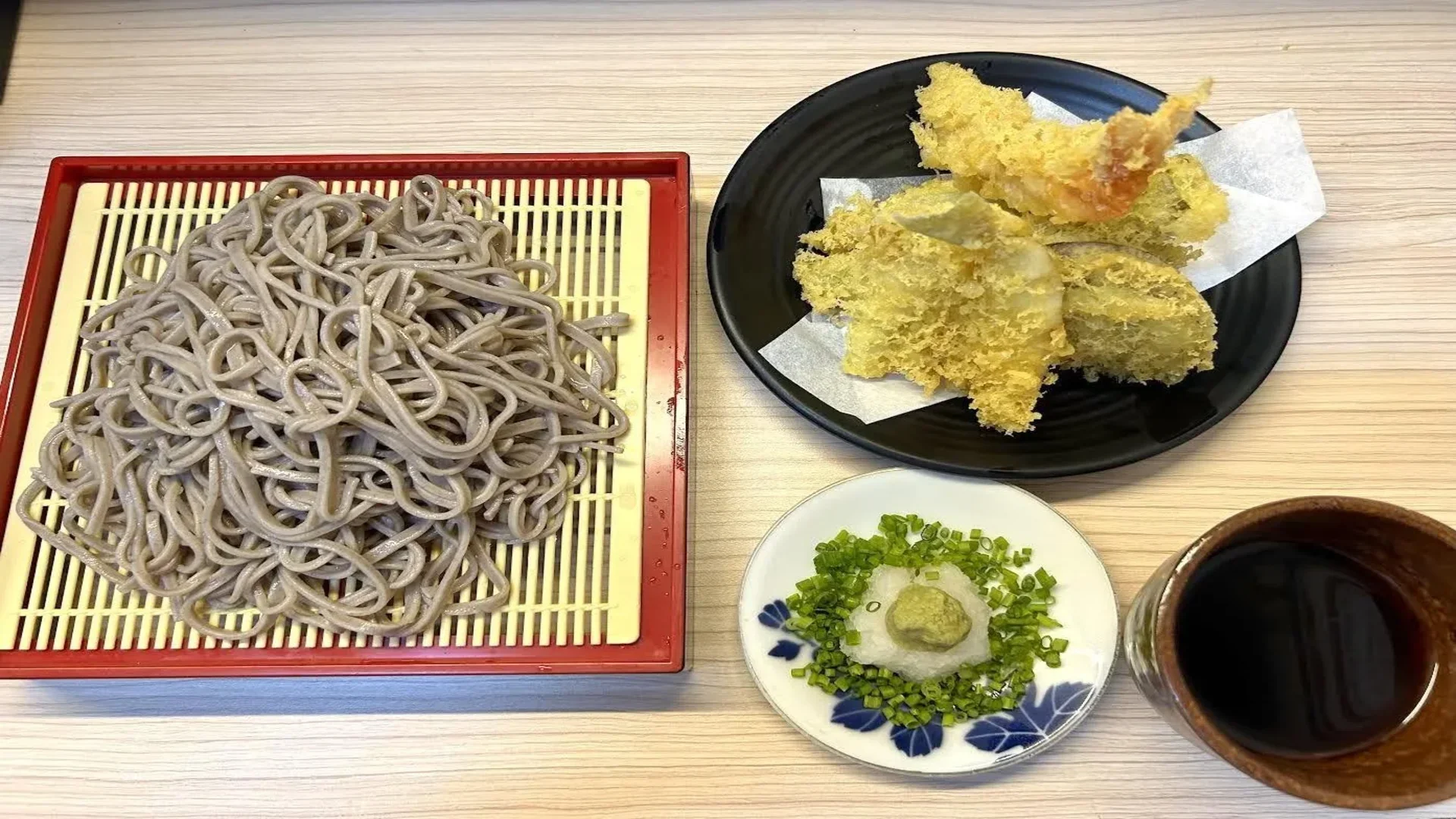 A plate of chilled soba noodles served with assorted tempura, green onions, grated daikon with wasabi, and dipping sauce on a wooden table.