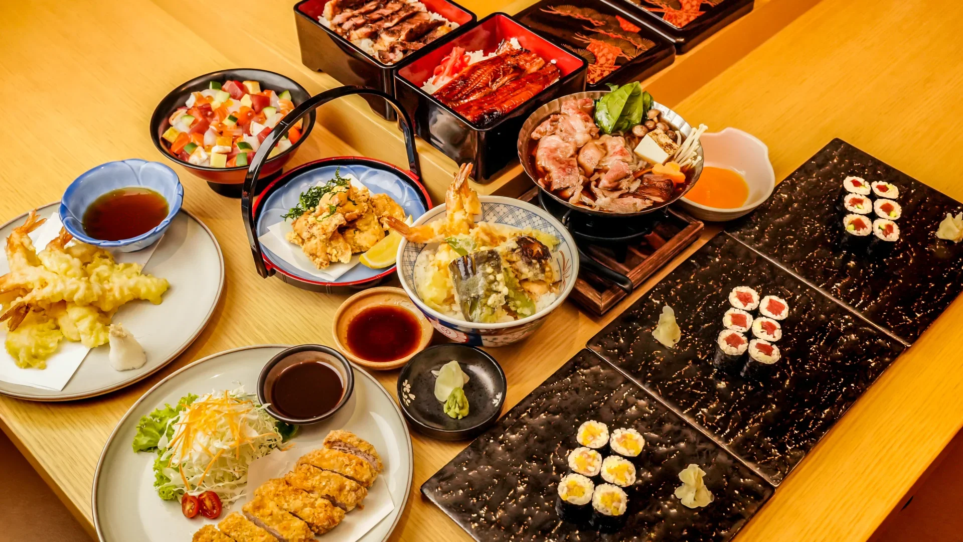 Japanese feast featuring sushi rolls, shrimp and vegetable tempura, fried dishes, sashimi, and hot pot arranged across a dining table.