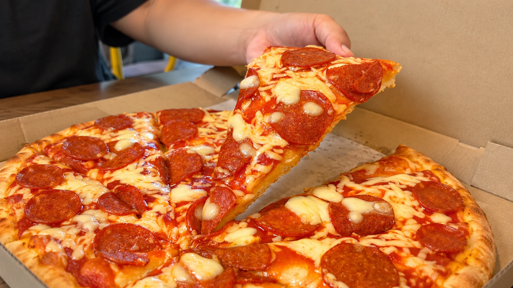 Hand lifting a cheesy pepperoni pizza slice from a freshly baked pizza inside a takeaway box.