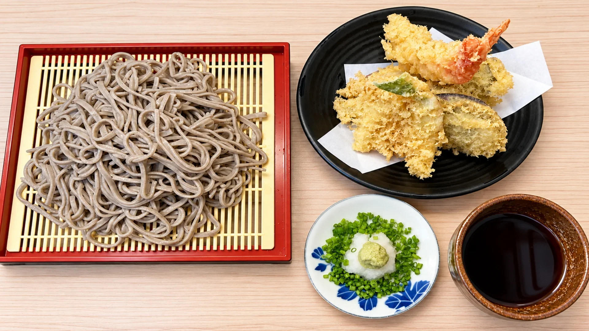 A plate of chilled soba noodles served with assorted tempura, green onions, grated daikon with wasabi, and dipping sauce on a wooden table.