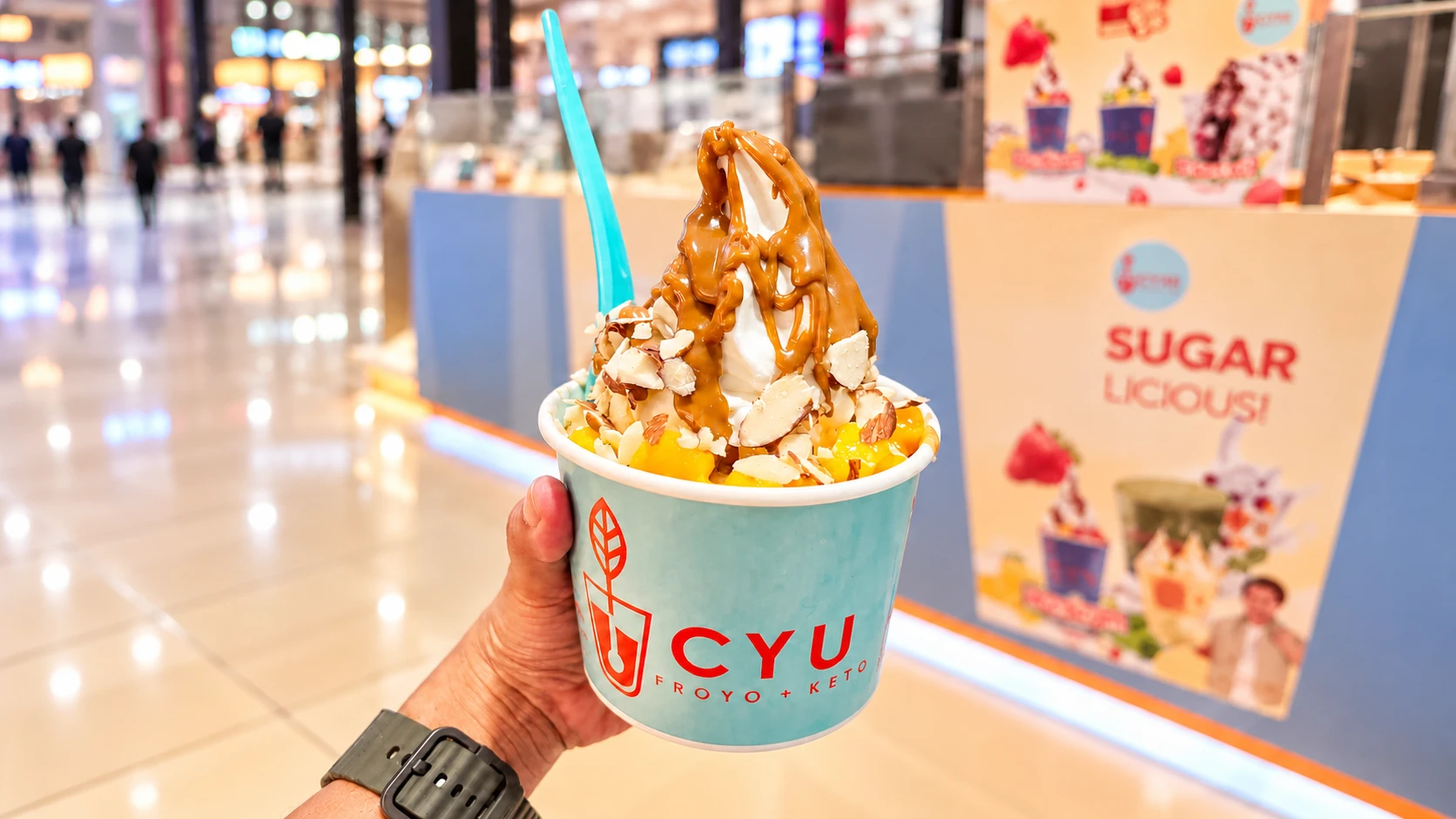 CYU Froyo + Keto frozen yogurt topped with caramel sauce, sliced almonds, and mango, held in front of the dessert kiosk.